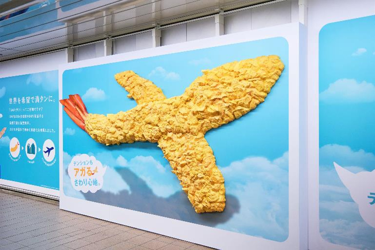 コスモエネルギーホールディングス、超サフサフな「飛行機型巨大エビ天」をモチーフにした屋外広告を新宿駅で掲出開始
